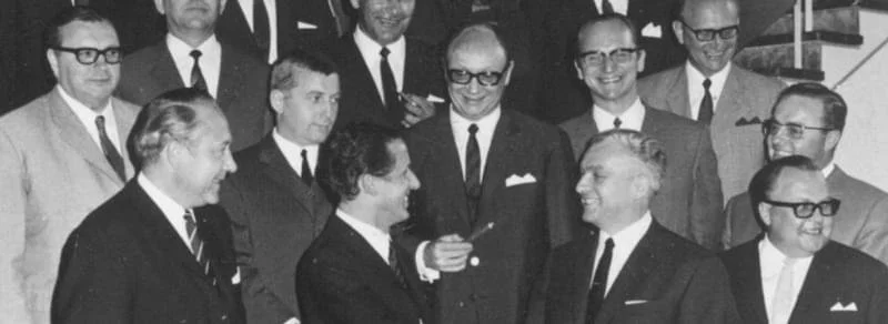 Mehrere Männer in Anzügen, einige mit Brillen, die sich in Gesprächen auf einer Treppe versammeln, in Schwarz-Weiß