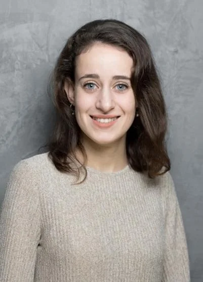 Ein junges Mädchen mit braunen, lockigen Haaren, blauen Augen und einem hellen Pullover vor einer grauen Wand.