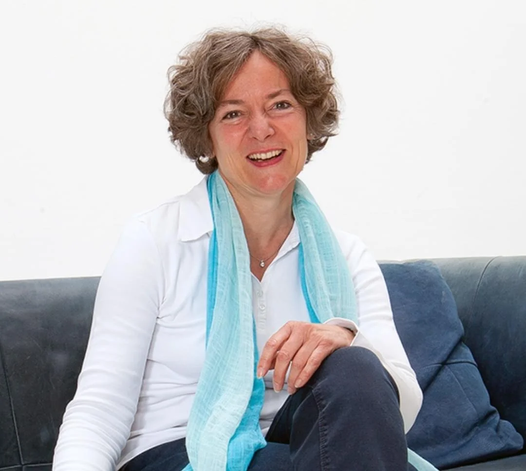 Eine ältere Frau mit lockigem braunem Haar sitzt auf einem Sofa, trägt ein weißes Hemd und eine helle blaue Stola, lächelt freundlich, vor einem weißen Hintergrund.