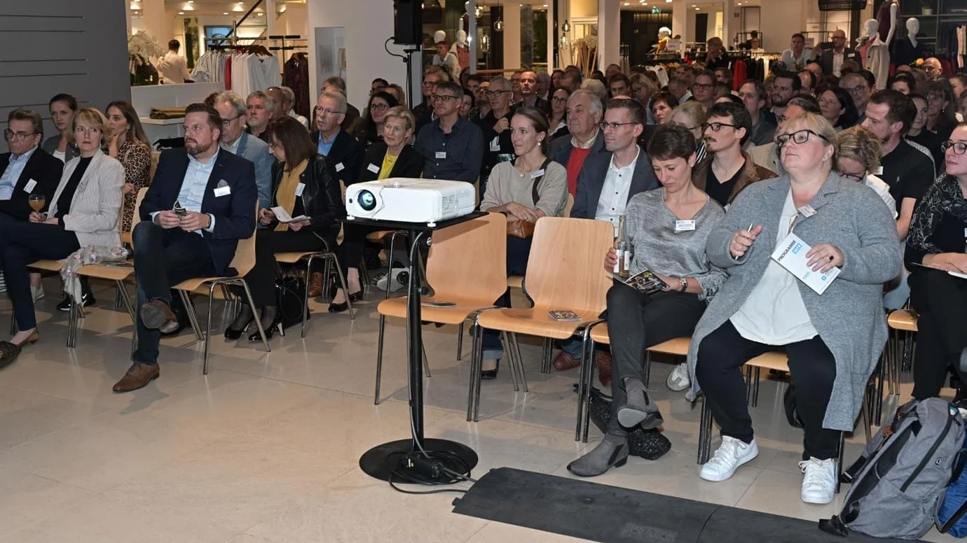 Menschen sitzen in einem Verkaufsraum und schauen einen Vortrag, der von einem Projektor auf eine Wand oder Leinwand übertragen wird.