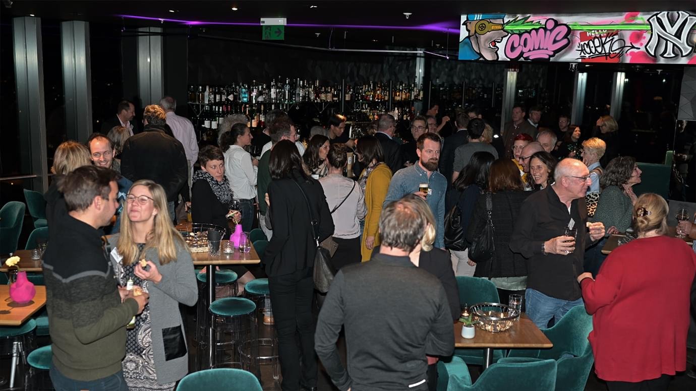 Menschen feiern in einer Bar, einige trinken, andere unterhalten sich, im Hintergrund eine Bar mit Flaschen, an der Decke bunte Graffiti-Kunst.