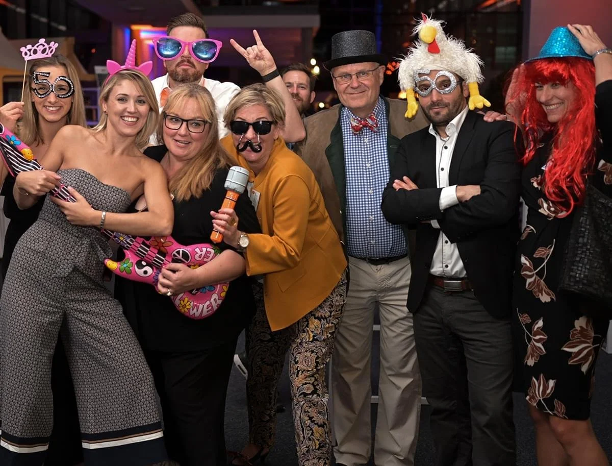 Menschen in Partykostümen und -accessoires bei einer Feier, einige tragen Hüte und lustige Brillen, lachen und posieren für das Foto.