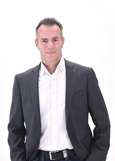 Ein Mann in Anzug und weißem Hemd, vor hellem Hintergrund, posiert für ein Porträt.