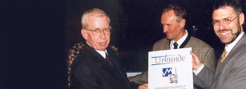 Drei Männer bei einer formellen Veranstaltung, einer hält eine Urkunde mit dem Titel 'Urkunde' und einem Bild, die anderen zwei lächeln.