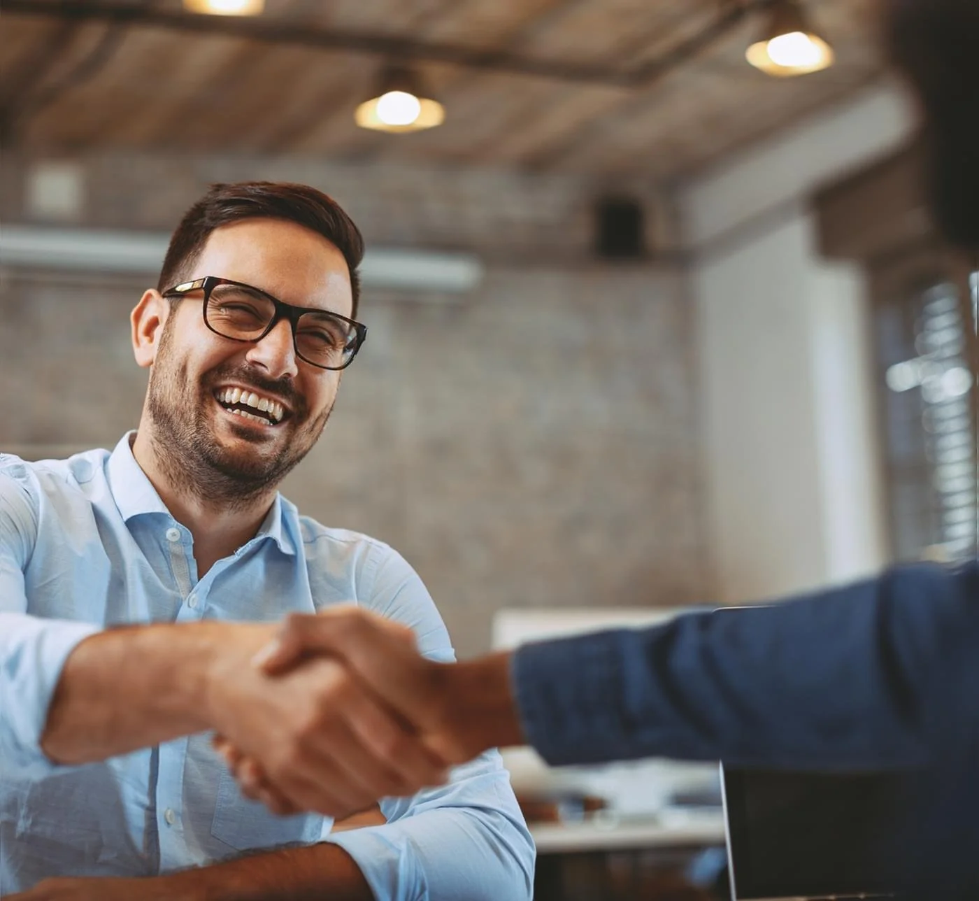 Zwei Männer schütteln sich die Hände und lachen. Sie sitzen in einem modernen Büro mit Holzbalkendecke und großen Fenstern.