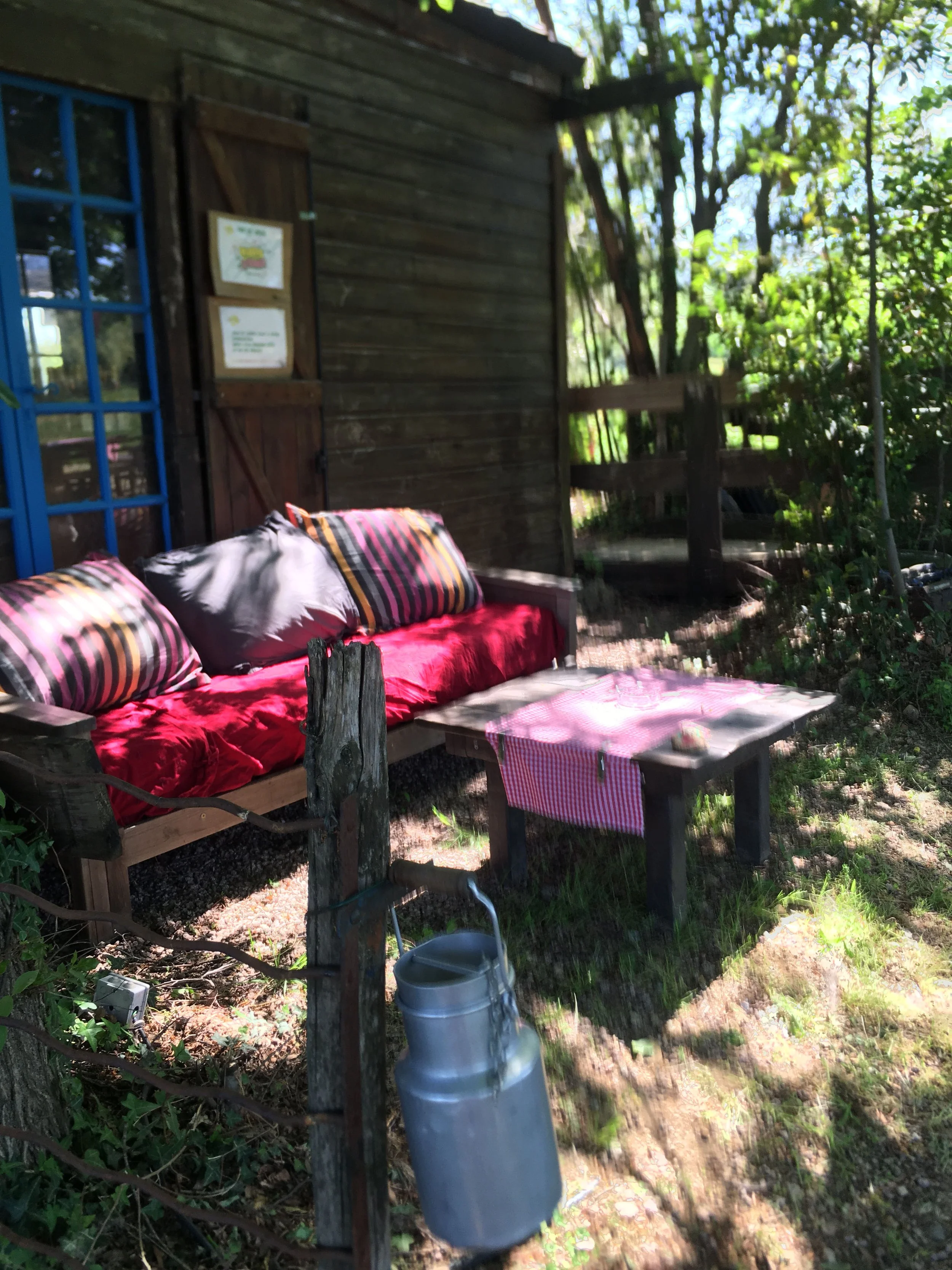 Une terrasse en plein air avec un canapé en bois, des coussins rayés et un coussin gris, une petite table recouverte d'un torchon à carreaux rouges et blancs, un mur en bois avec des fenêtres bleues, entourée de végétation abondante.