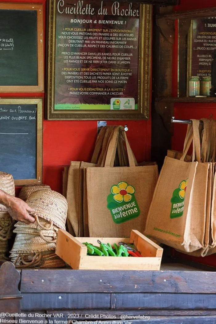Sac de courses en jute avec le logo « Bienvenue à la ferme » dans une boutique à la cueillette du rocher Présence d'outils de jardinage, paniers en osier et tableau d'affichage.