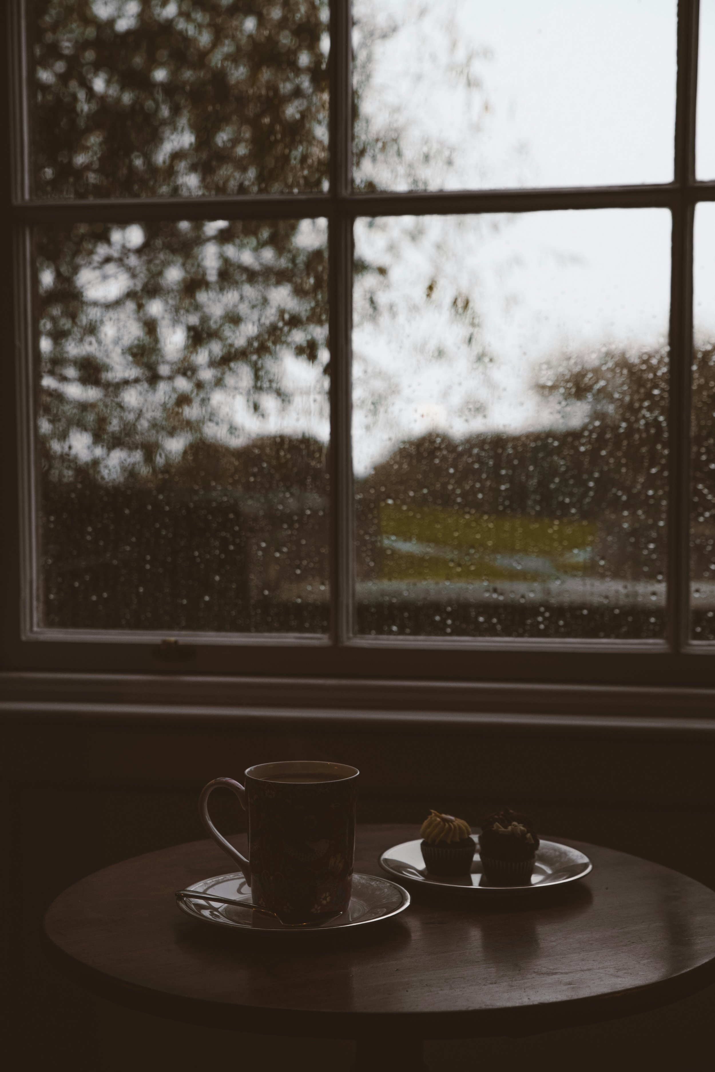 A dark wooden table with a coffee mug, a small spoon, and two cupcakes on white plates inside a room. Behind the window, raindrops are visible on the glass, with blurred trees and cloudy sky outside.