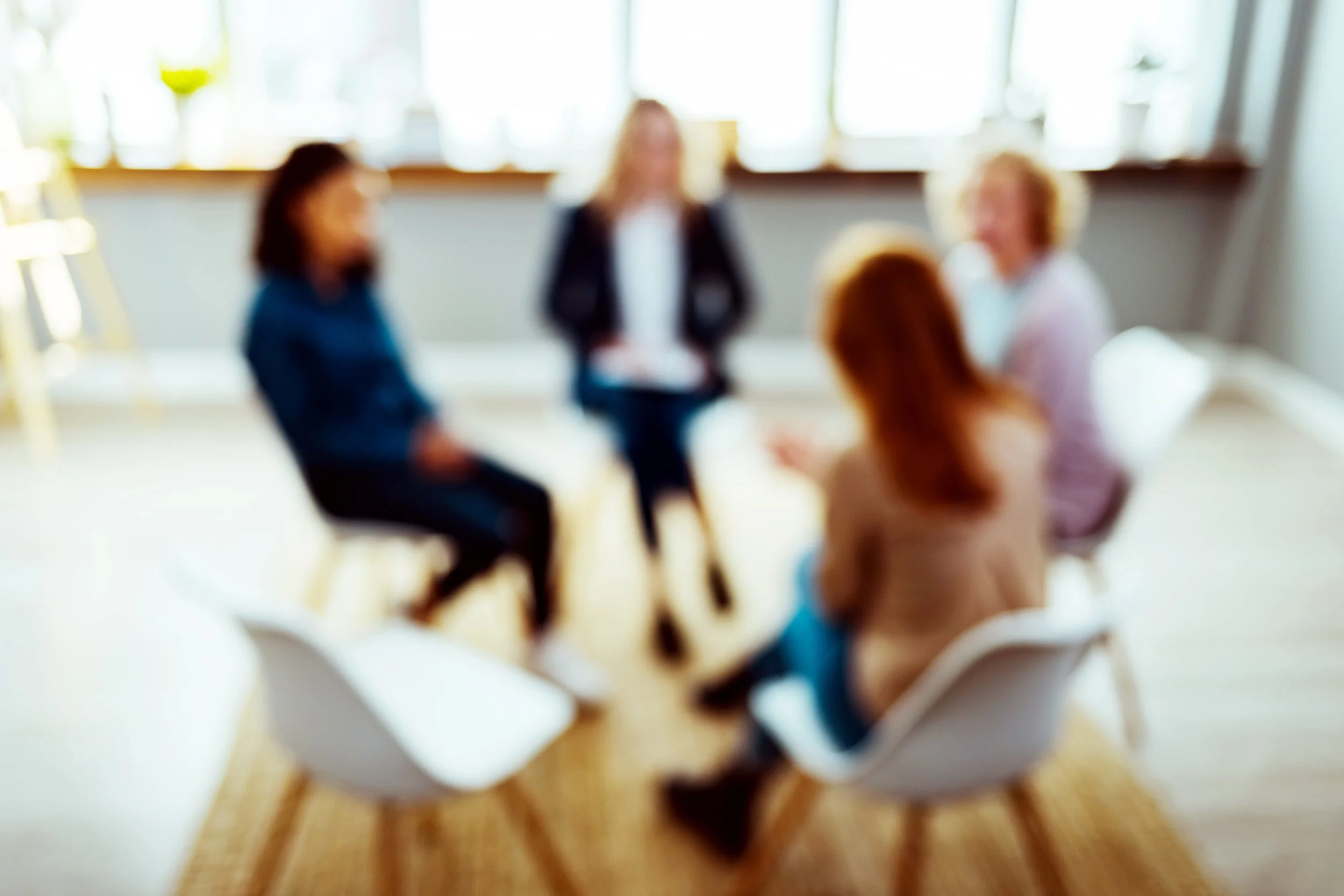 Five women in a support group circle having a discussion in a bright room with large windows discussing relational trauma, anxiety, and self-trust.