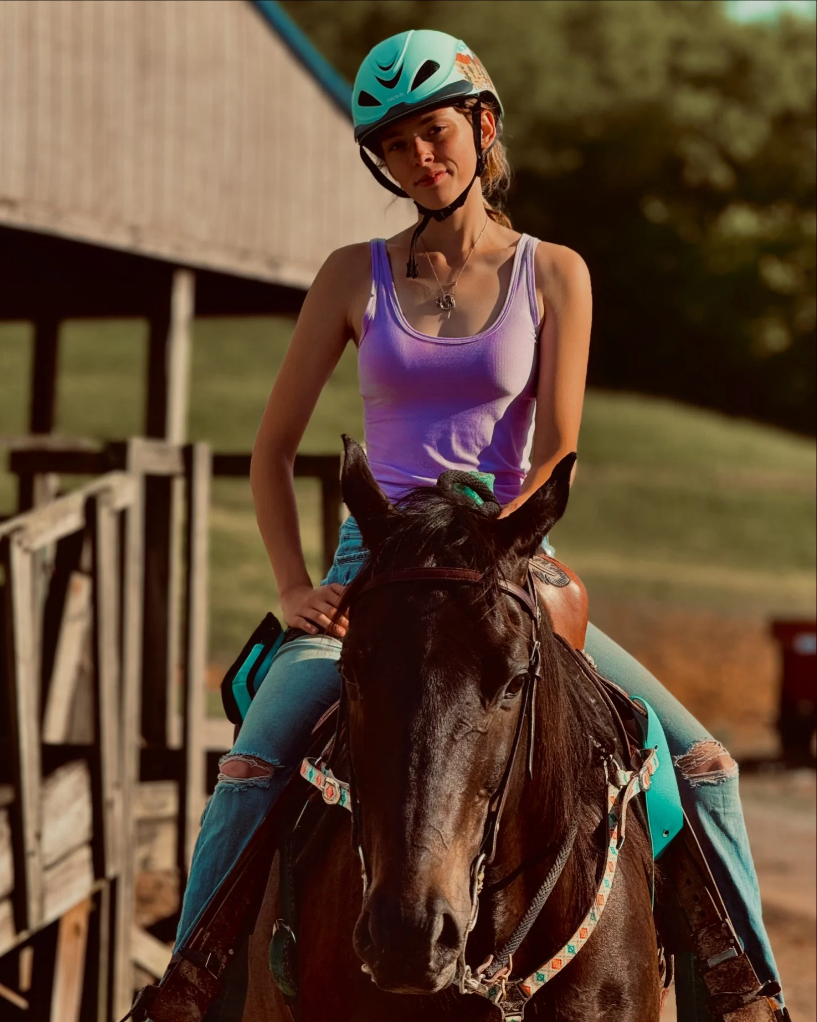 A young woman riding a horse outdoors. She is wearing a blue helmet, a purple tank top, ripped jeans, and is sitting on a brown horse with a colorful saddle.