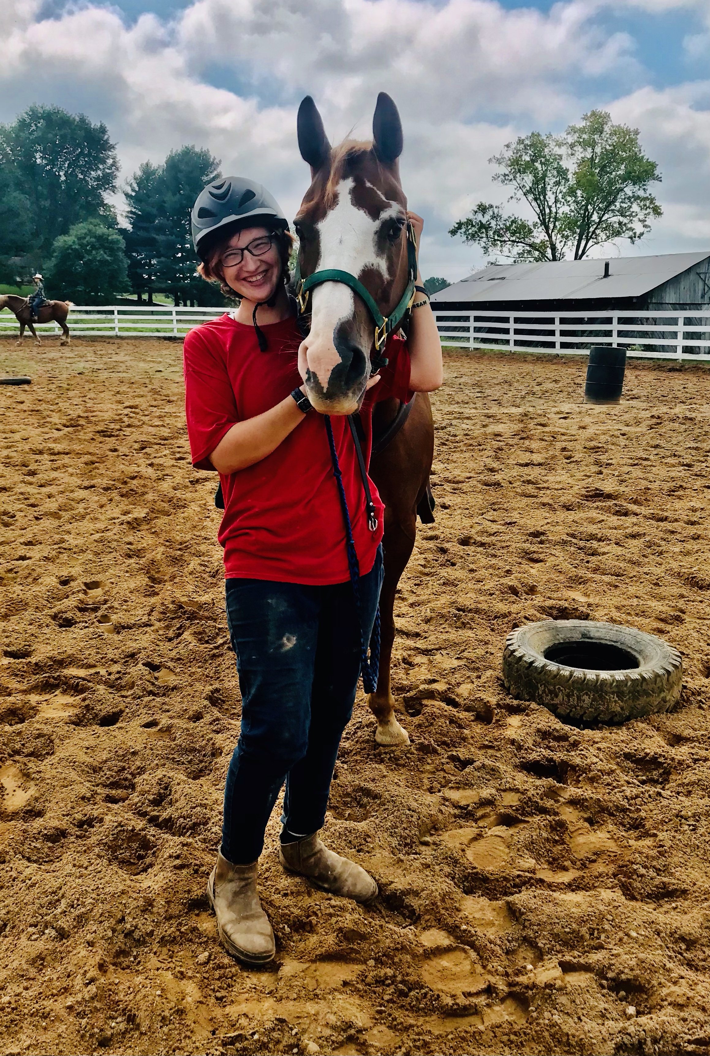 A smiling person in a red shirt and riding helmet hugging a brown and white horse in a sandy outdoor riding area.