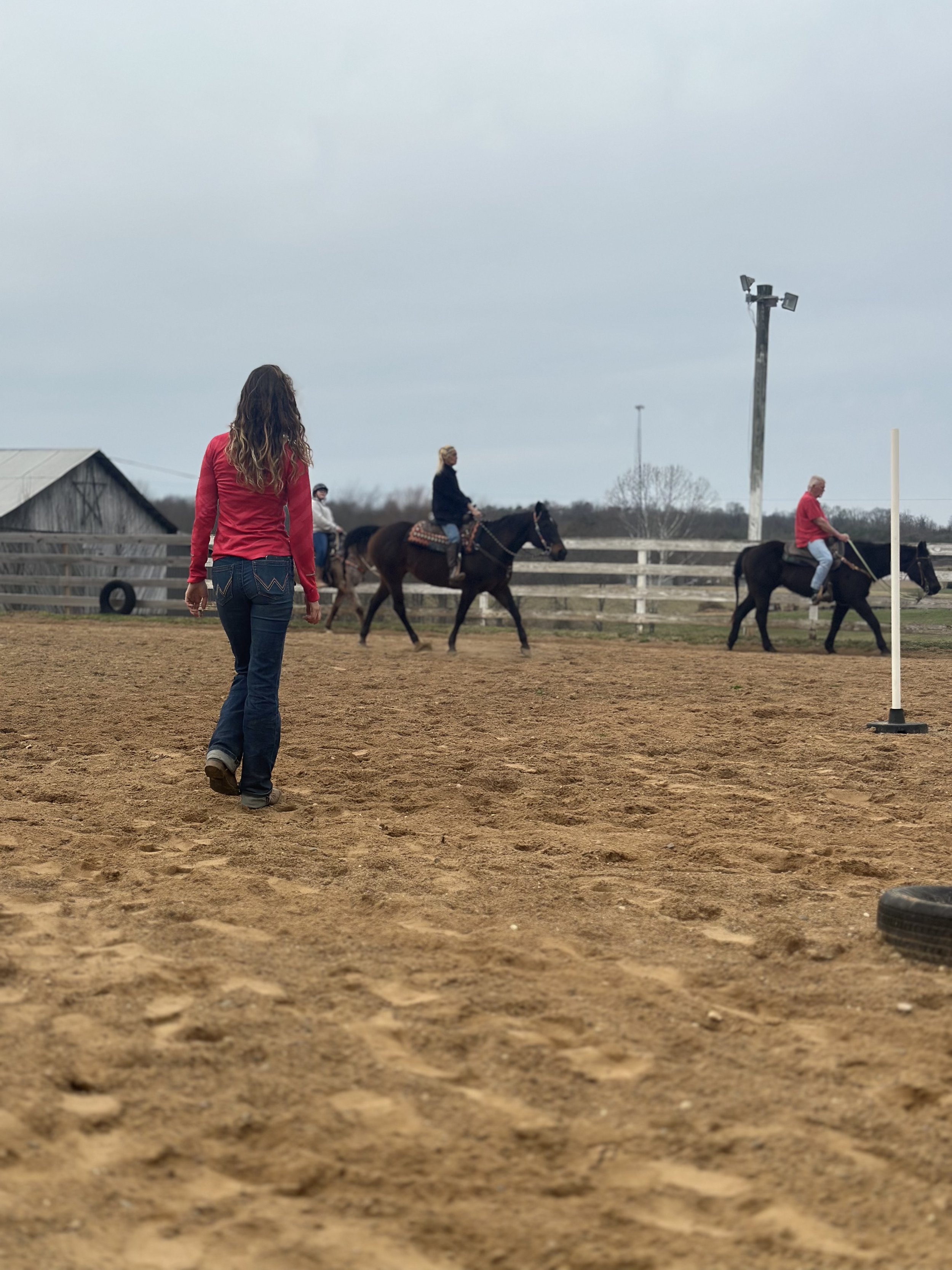A woman walks on a dirt outdoor riding arena past three people on horseback, with a fence, a barn, and cloudy sky in the background.