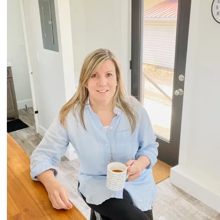 a woman with straight blonde hair sits near a glass door holding a mug of tea