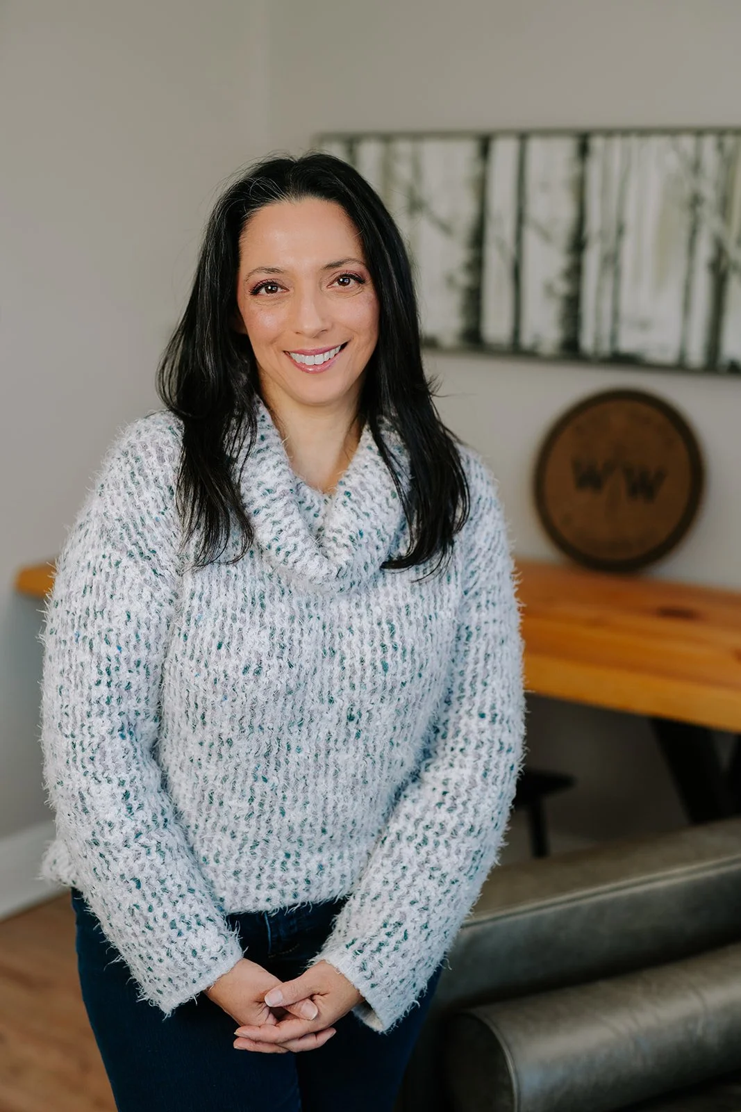 a woman with straight black hair wearing a blue and white sweater stands against a wall with a landscape photo