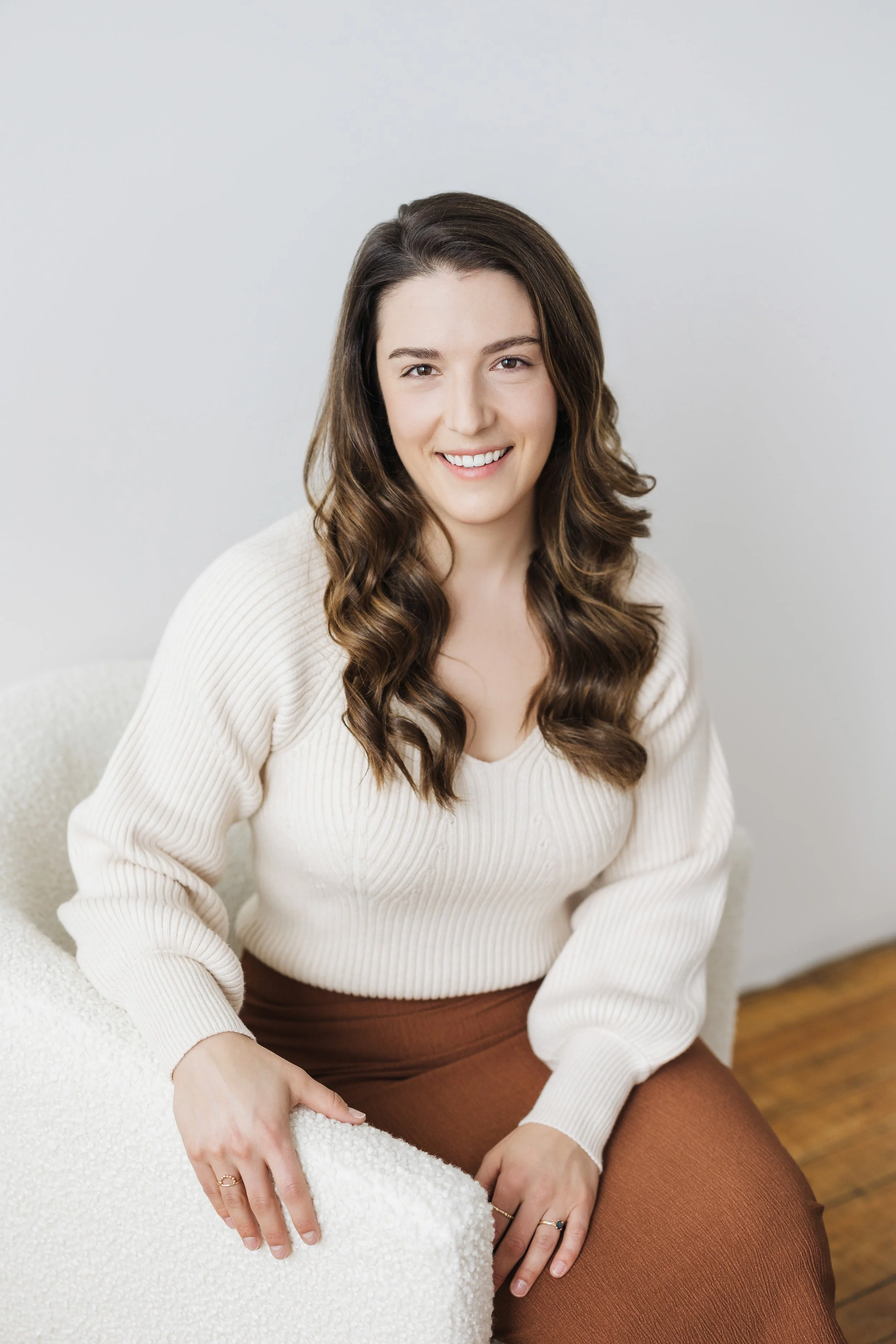 a woman with brown, curled hair in a cream sweater sits against a white wall
