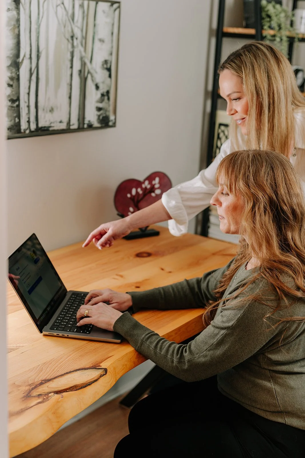 Two women working together on a laptop at a wooden desk in a cozy home office.