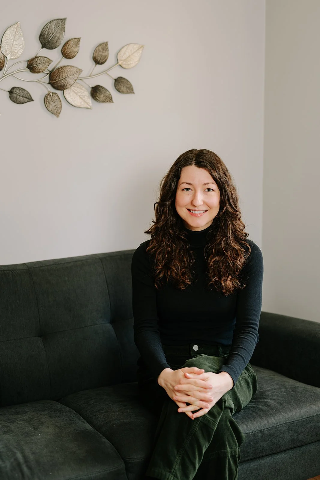 A woman with brown curly hair wearing a black turtleneck sweater sits on a dark couch with hands clasped on knee against white wall