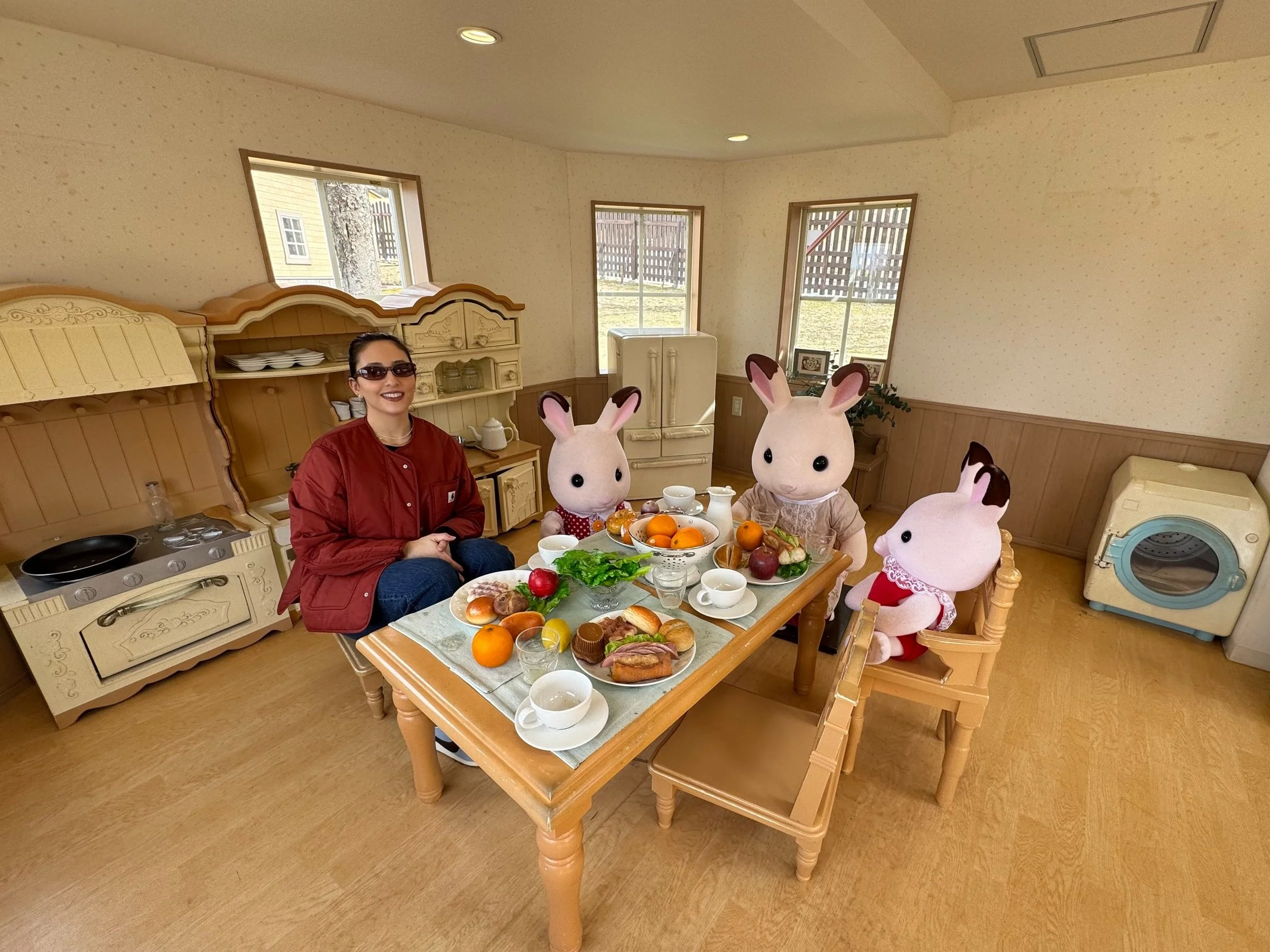 Woman sitting at a table with life-size Calico Critter figures at the Calico Critter Museum in Osaka, enjoying a meal in a cozy kitchen setting.