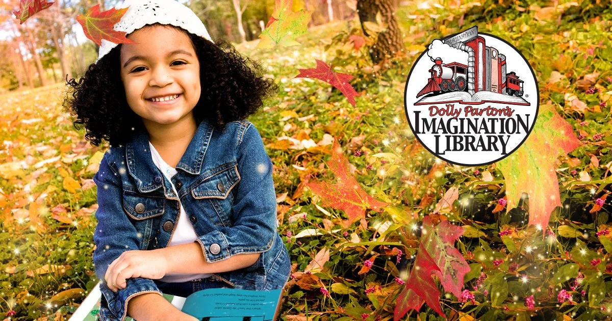 A young girl with curly hair smiling, sitting in a field with autumn leaves. She is wearing a denim jacket and holding a book. There is a logo for Dolly Parton’s Imagination Library in the background amongst fall foliage.