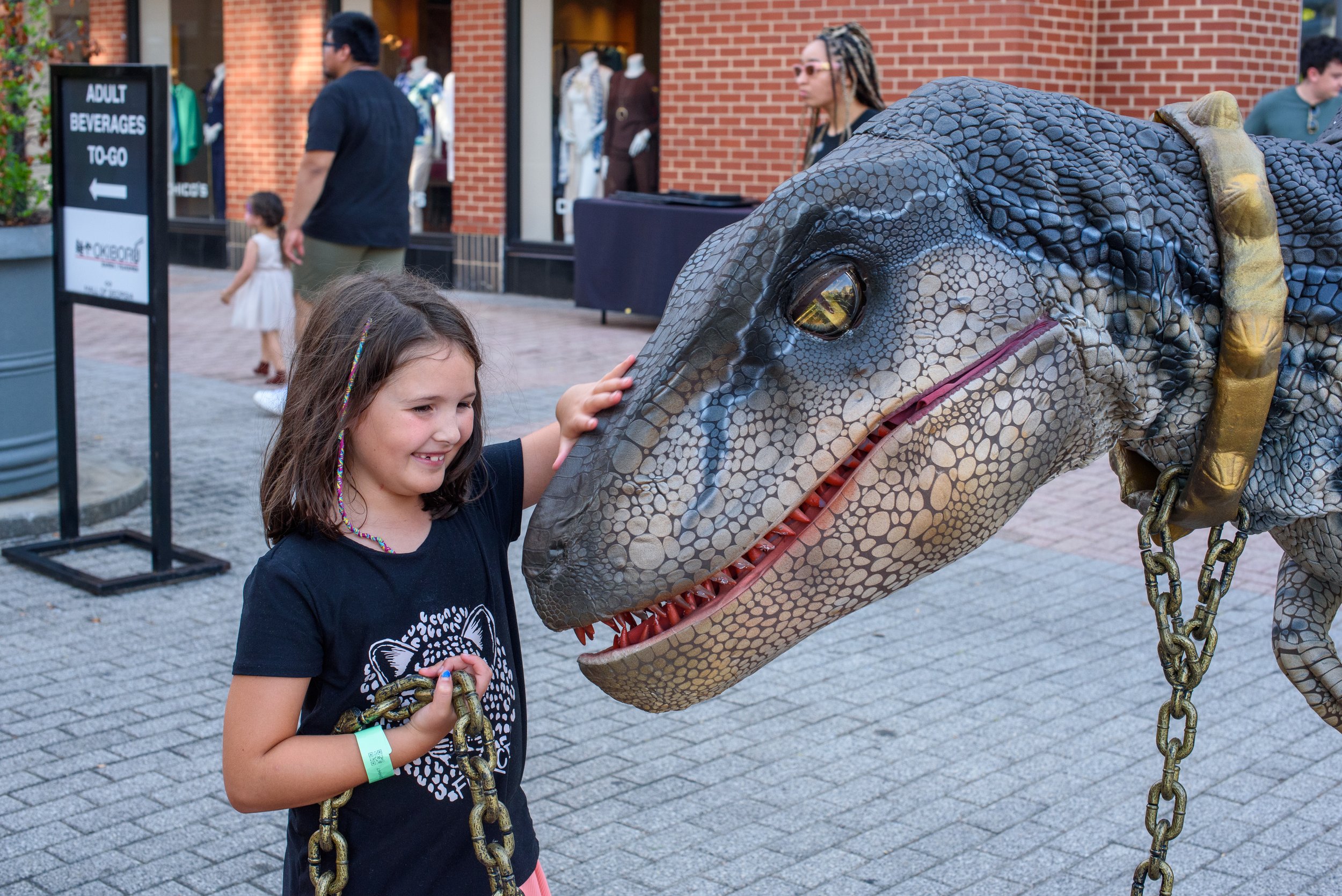 A young girl is petting a life-sized realistic dinosaur sculpture with textured gray and black scales. The dinosaur is chained to a post and stands outside a shopping area with brick buildings, while other people walk by in the background.