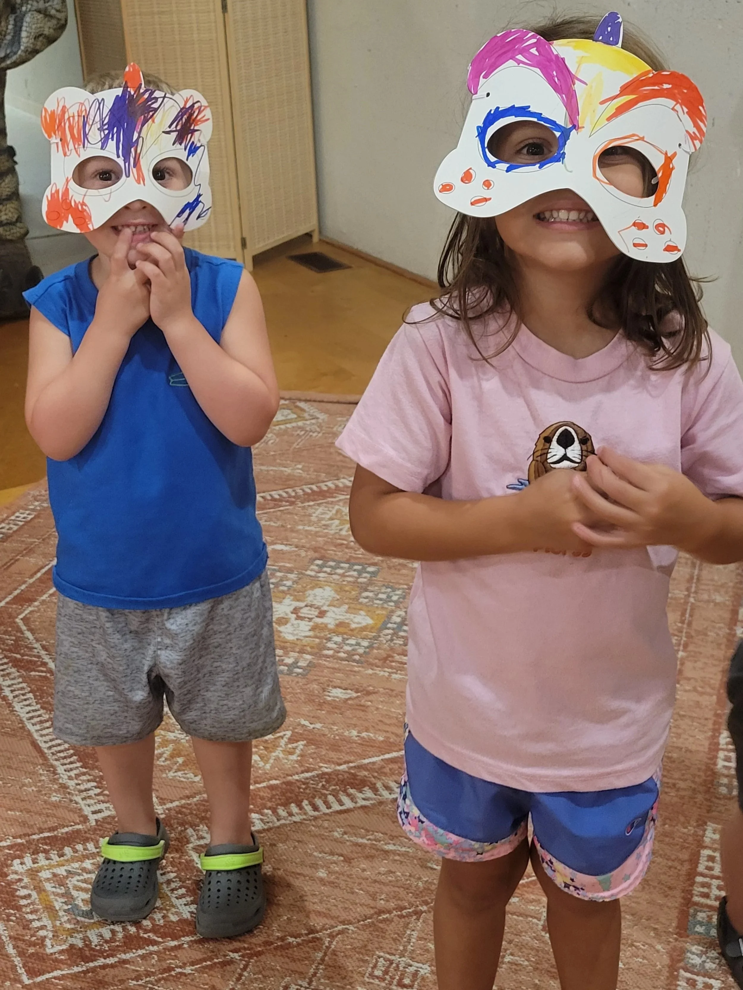 little boy in a blue shirt wearing a handmade colored dinosaur mask next to a little girl in a pink shirt wearing a dinosaur handmade dinosaur mask