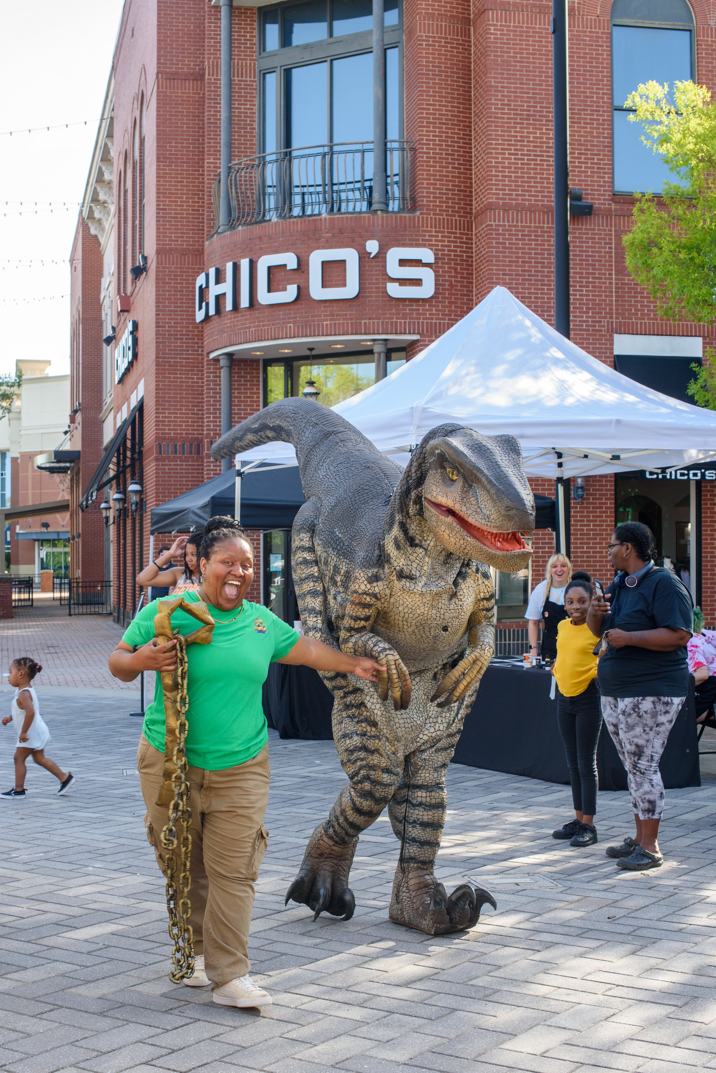 Woman holding the reigns of a gray Velociraptor dinosaur in front of a brick store front.