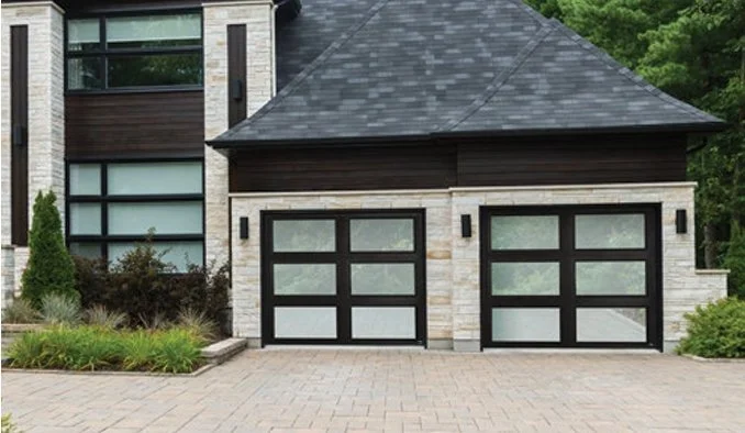 Modern house exterior with two black-framed glass garage doors, light brick facade, and paved driveway.