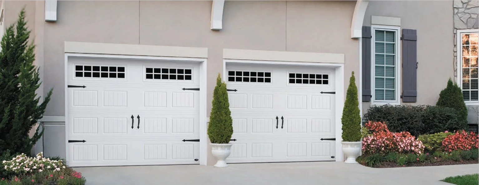 Two white garage doors with black hardware design elements, flanked by potted trees, in a modern residential setting. The garage is surrounded by trimmed hedges and flowering plants.