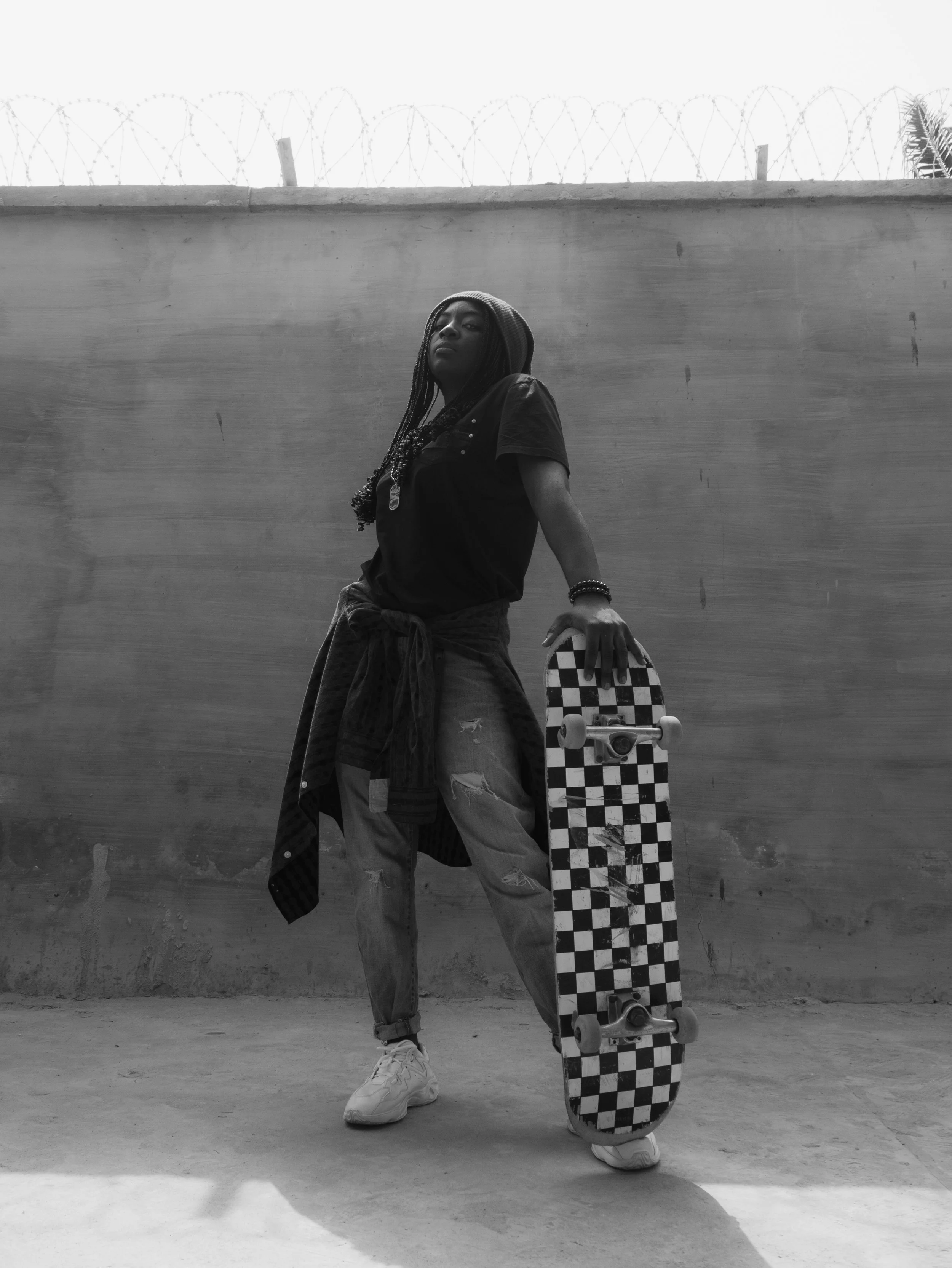 Joven con camiseta negra, pantalones rasgados y sudadera atada a la cintura, sosteniendo una patineta con diseño de cuadros en una pared de color claro, baranda de alambre de púa en el fondo.