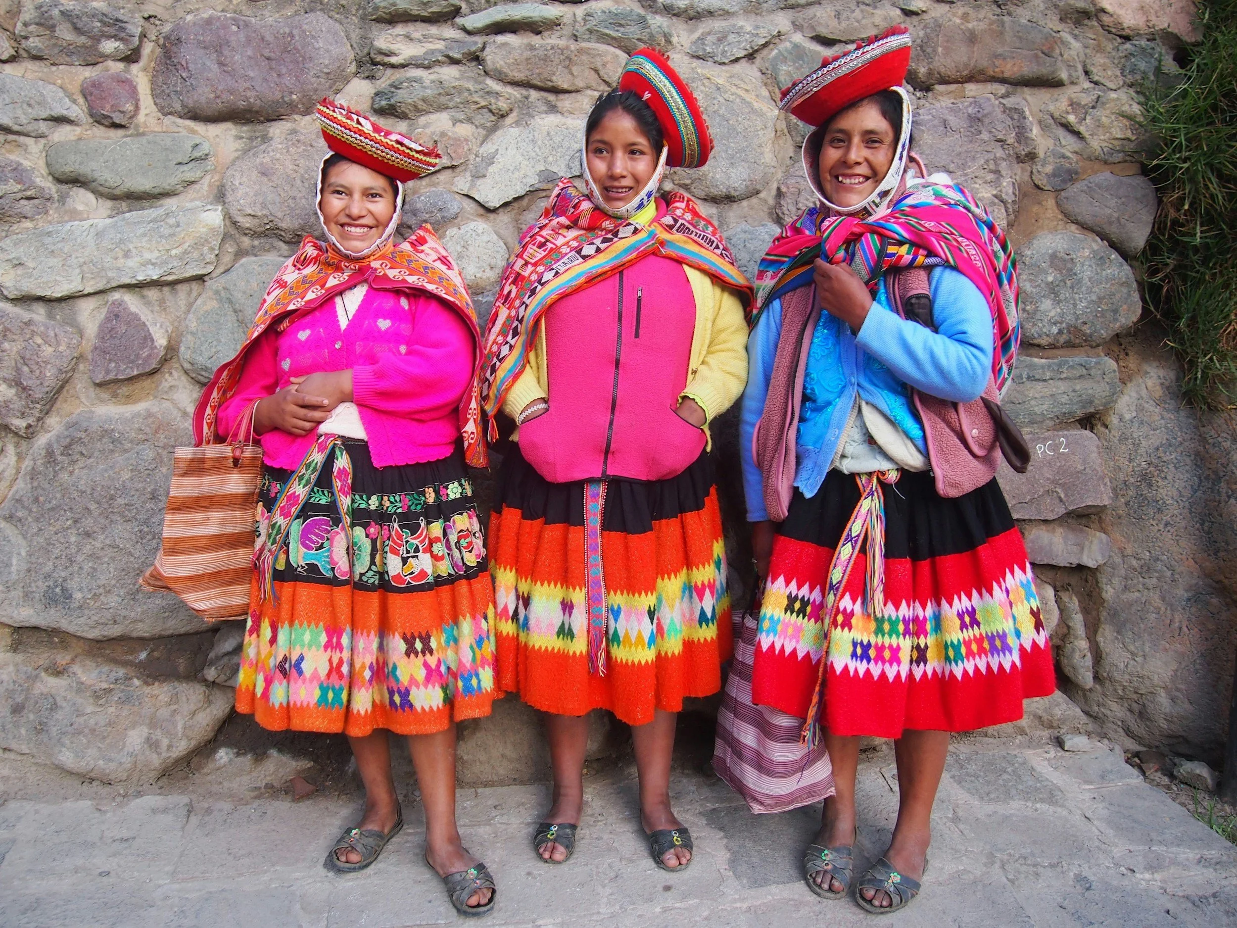 Three women wearing traditional colorful Andean clothing and hats, standing against a stone wall, smiling and posing for the camera.