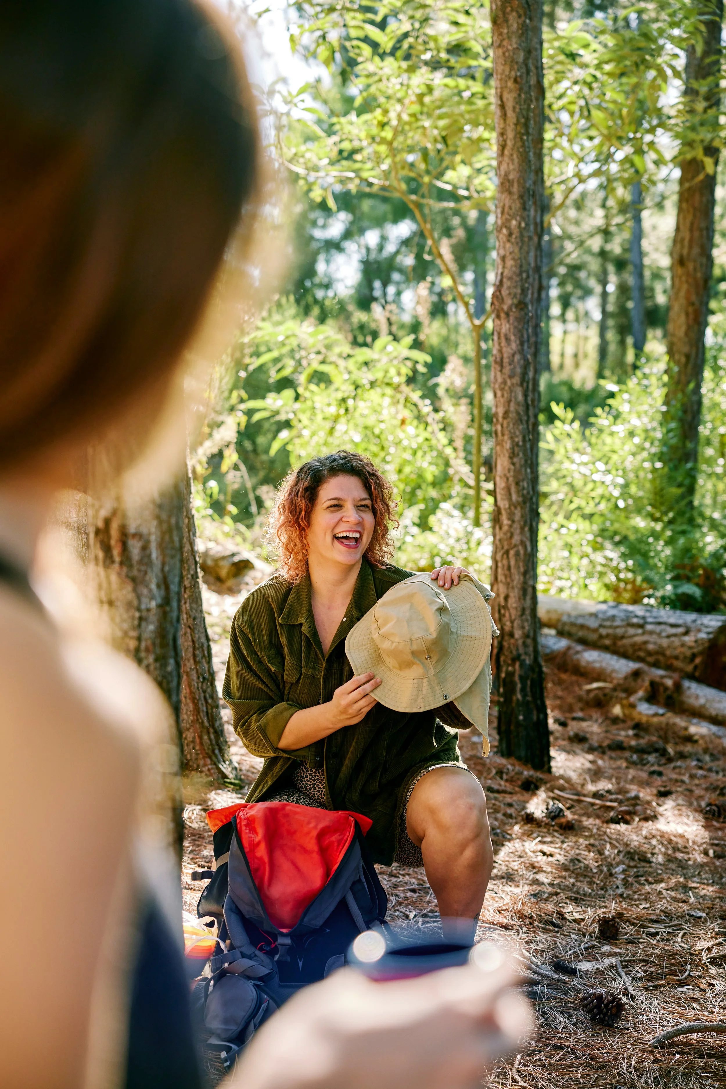 A woman with curly red hair sitting on the forest ground, smiling and holding a wide-brimmed hat, with a backpack nearby, in a wooded area with tall trees and sunlight filtering through the leaves.