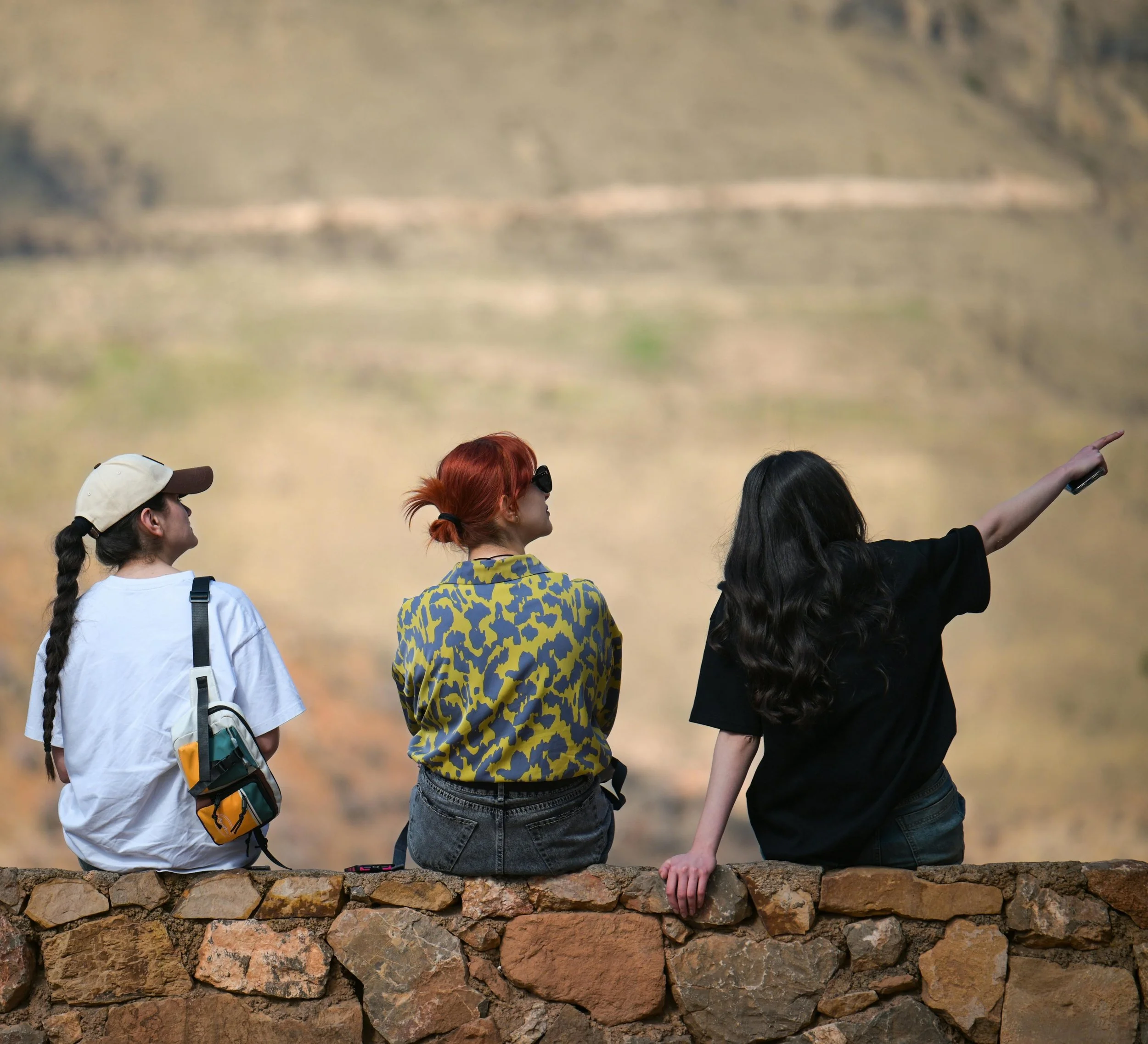 Three women sitting on a stone wall, observing a mountainous landscape, with one woman pointing towards the distance.