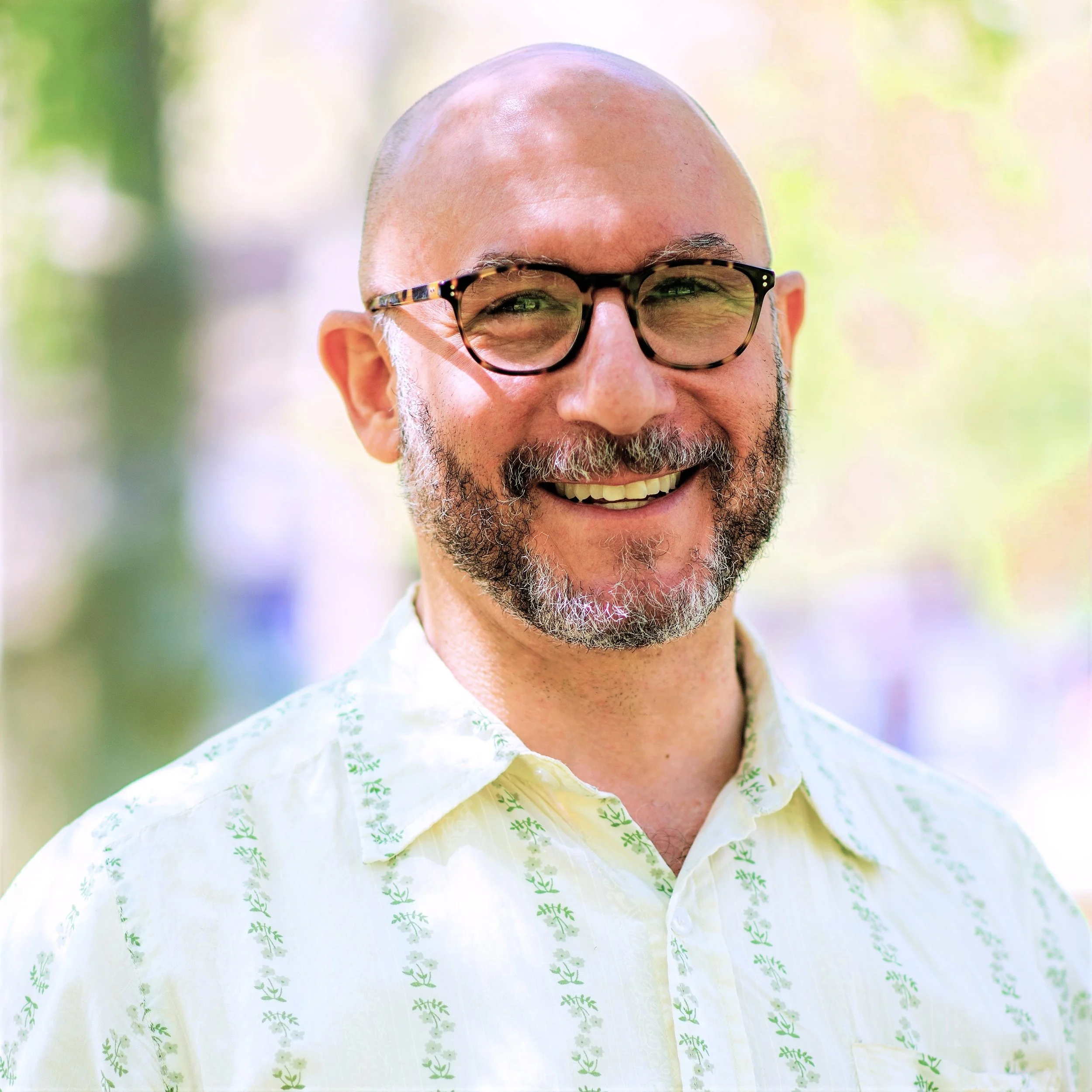 Man smiling outdoors, wearing glasses and a patterned shirt