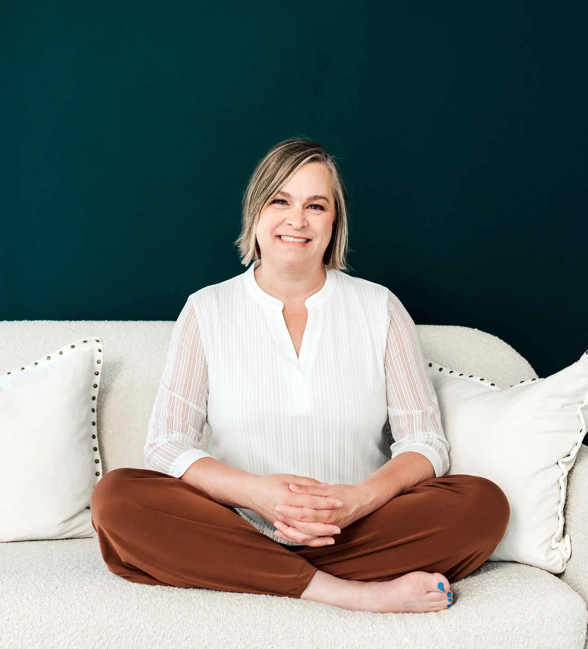 A woman with blonde hair sitting cross-legged on a white carpet with her hands resting on her knee. She is smiling and wearing a white top and brown pants. Behind her are two white cushions on a couch, against a dark teal wall.