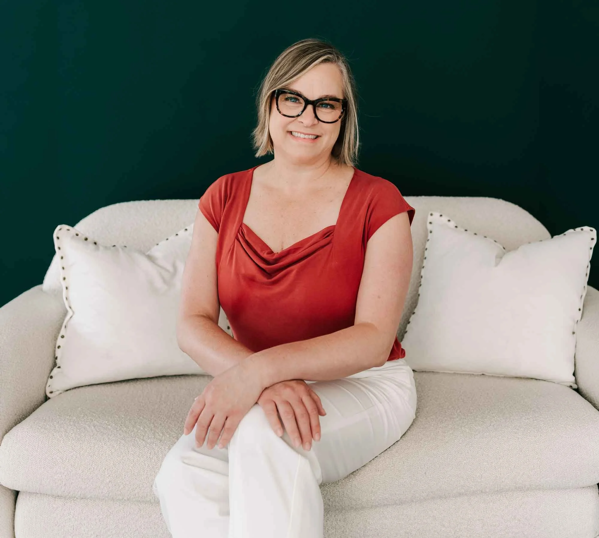 A woman with short blond hair, wearing glasses, a red top, and white pants, sitting on a light-colored sofa with white pillows against a dark green wall.