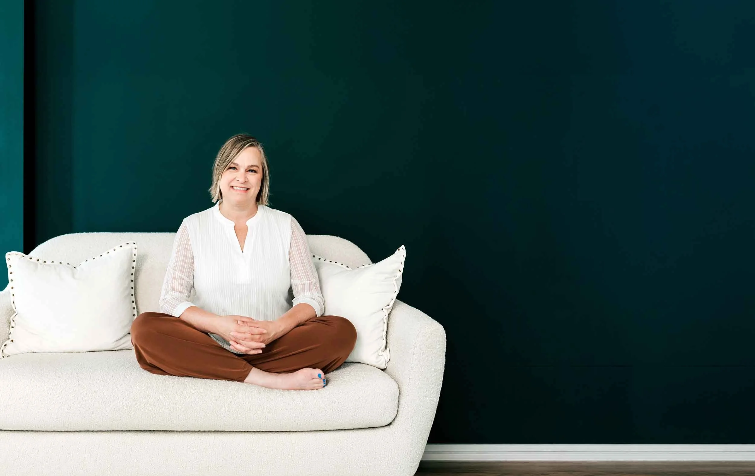 A woman with short blonde hair, wearing a white blouse and brown pants, sitting cross-legged on a light-colored sofa, smiling with a dark green wall in the background.
