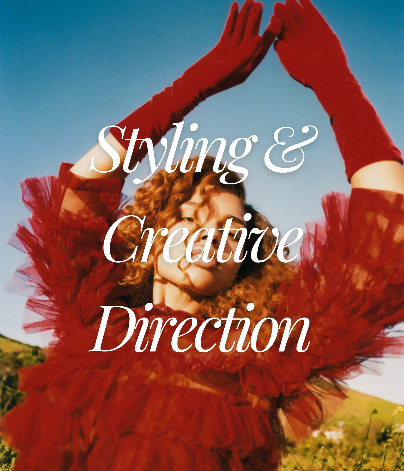 A woman with curly red hair wearing a red tulle dress and long red gloves, standing outdoors against a blue sky, with the words 'Styling & Creative Direction' overlaid.