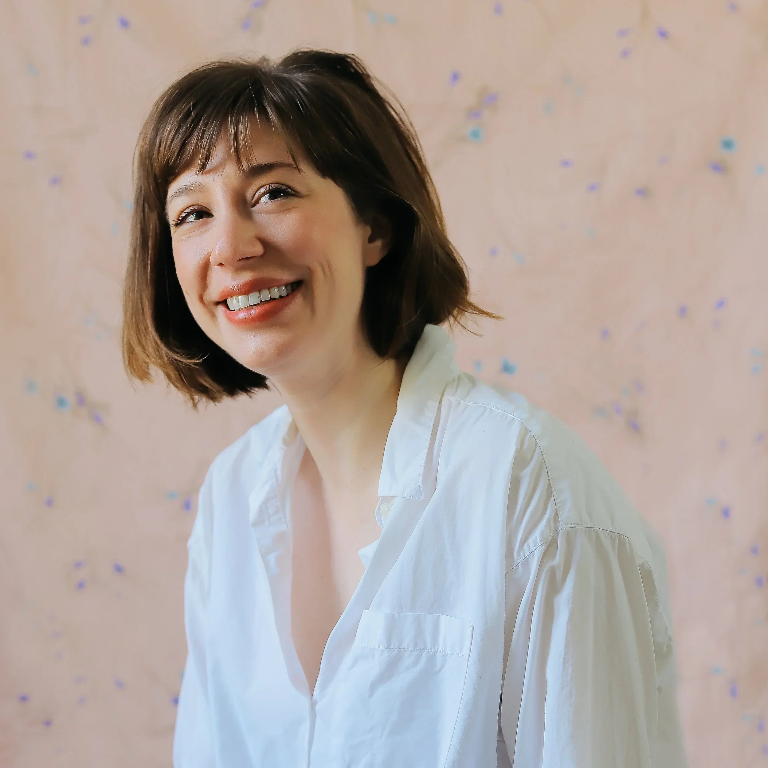 Director Annie Tippe. A woman with short brown hair smiling, wearing a white shirt, with a blurred pink background decorated with small blue and purple confetti.