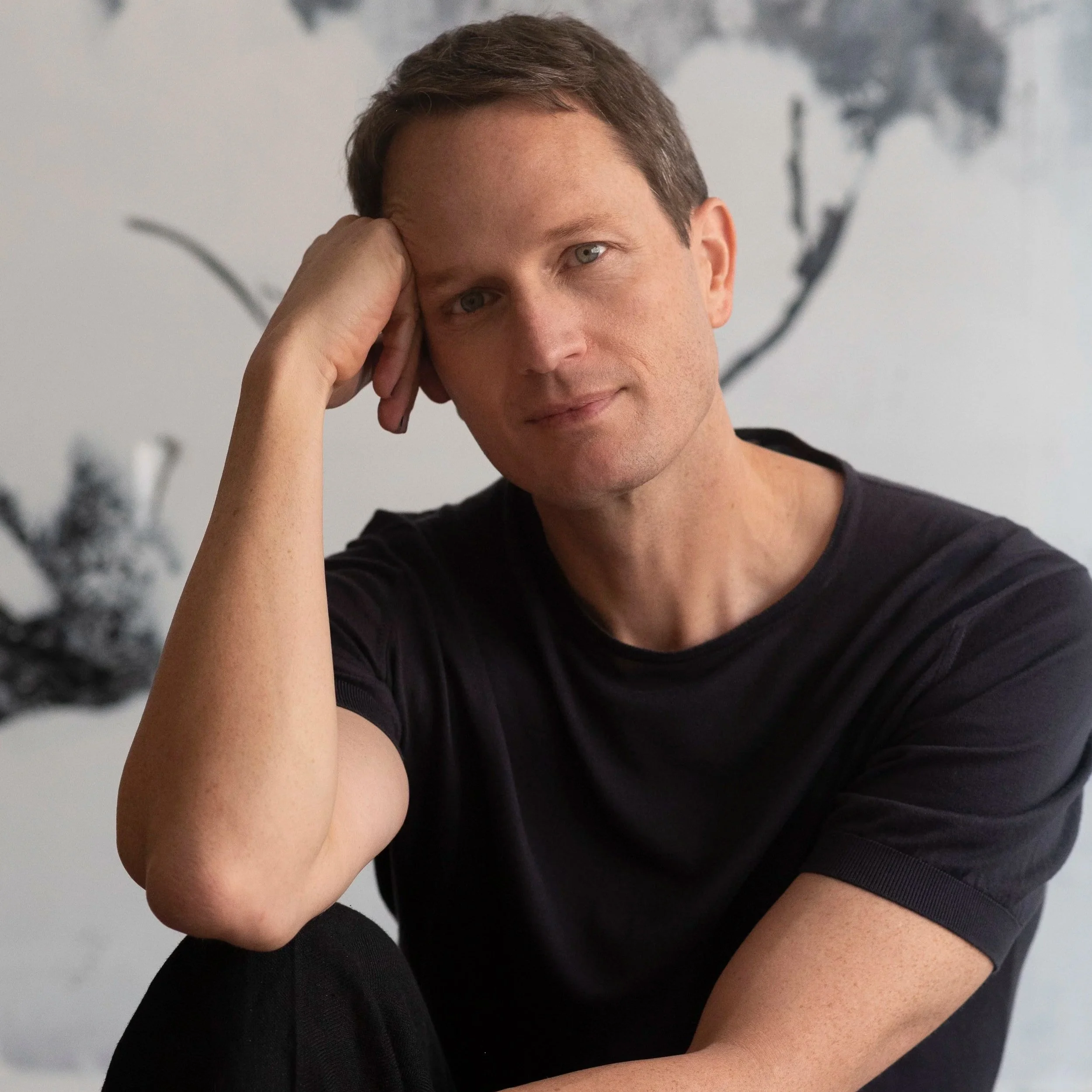 Song co-writer Todd Almond. A cis man with short brown hair, wearing a black T-shirt, resting his head on his hand and looking at the camera against a blurred abstract background.