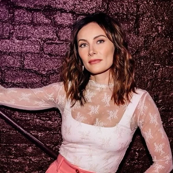 Woman with wavy brown hair, wearing a white lace top with sheer long sleeves and pink pants, standing against a textured dark background.