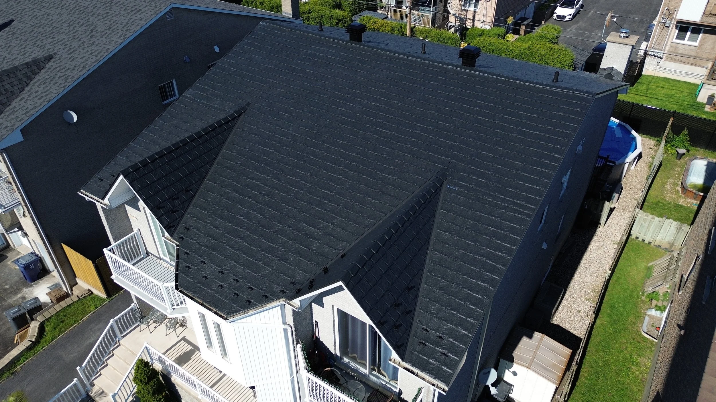 Vue aérienne d'une maison avec un toit en ardoise noire, comprenant des zones en pente et une petite toiture en tôle noire sur la façade, avec un balcon blanc et un espace extérieur pavé attenant.