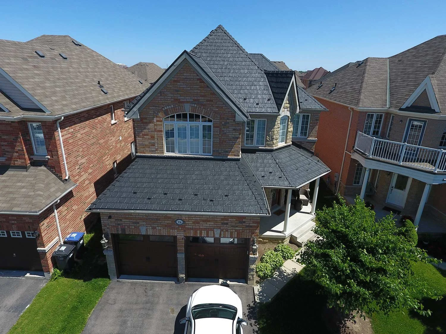 Vue aérienne d'une maison en briques avec garage et voiture garée devant, entourée d'autres maisons similaires, sous un ciel clair.