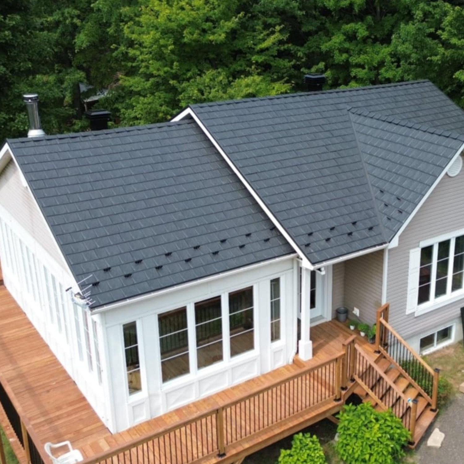 Maison avec toiture en tuiles noires, terrasse en bois, fenêtres large, et entourée de verdure.