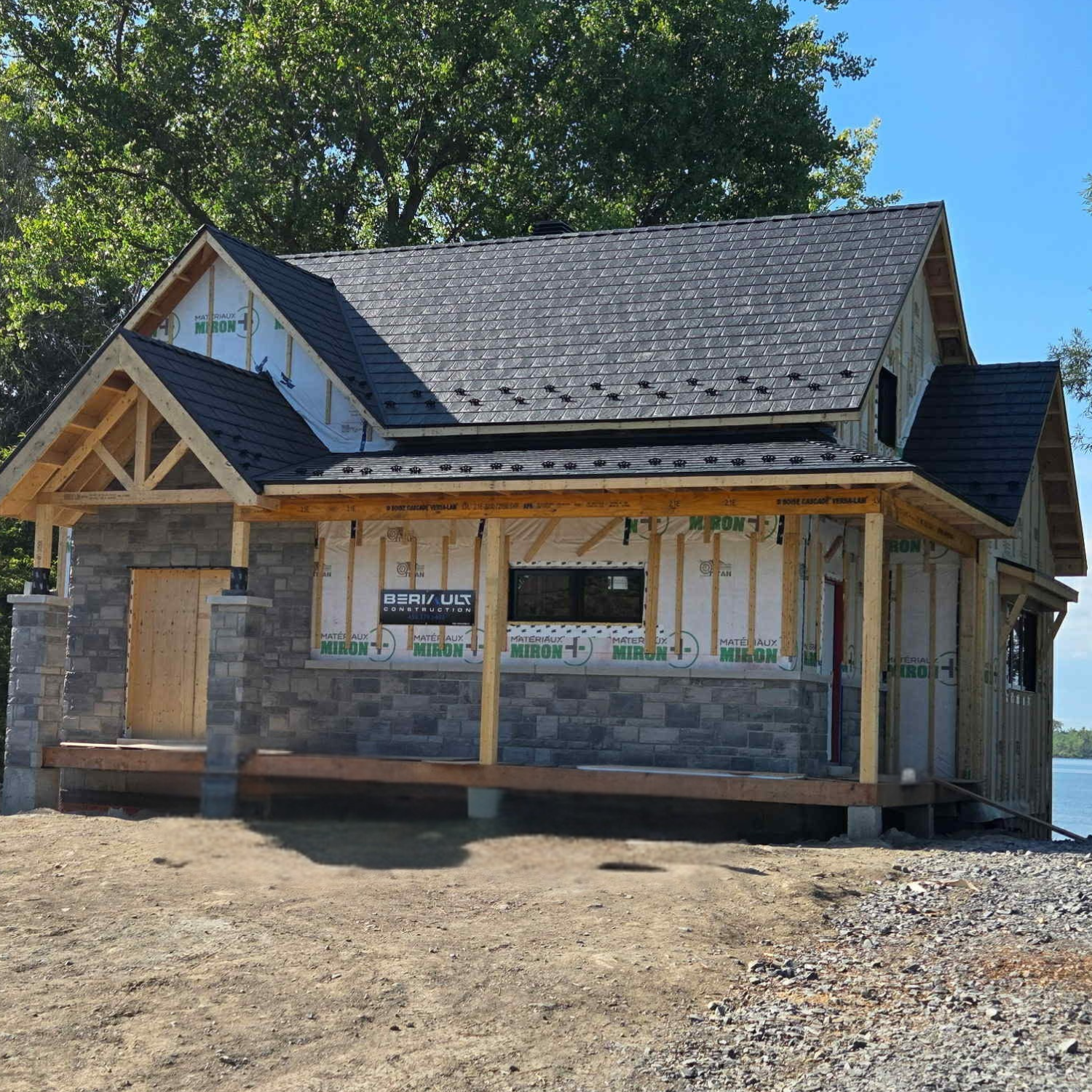 Une maison en construction avec une structure en bois, murs en pierre, fenêtres et toiture partiellement terminées.