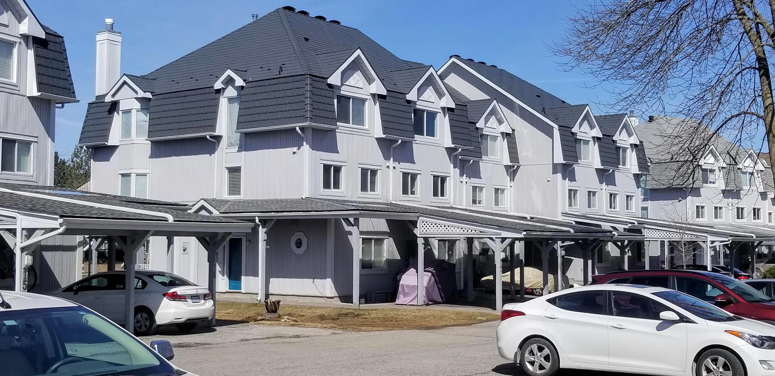Immeubles en béton blanc avec plusieurs étages, toits en pente noire, parking avec voitures blanc, rouge et gris, arbre sans feuilles en premier plan à droite, ciel bleu clair.