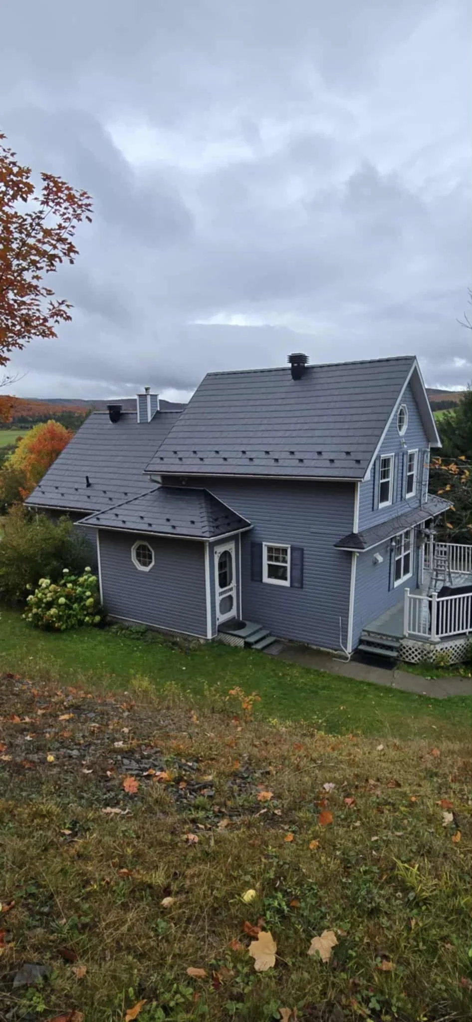Maison bleue à deux étages avec toit en tuiles grises, chêne avec feuilles d'automne, et ciel nuageux.