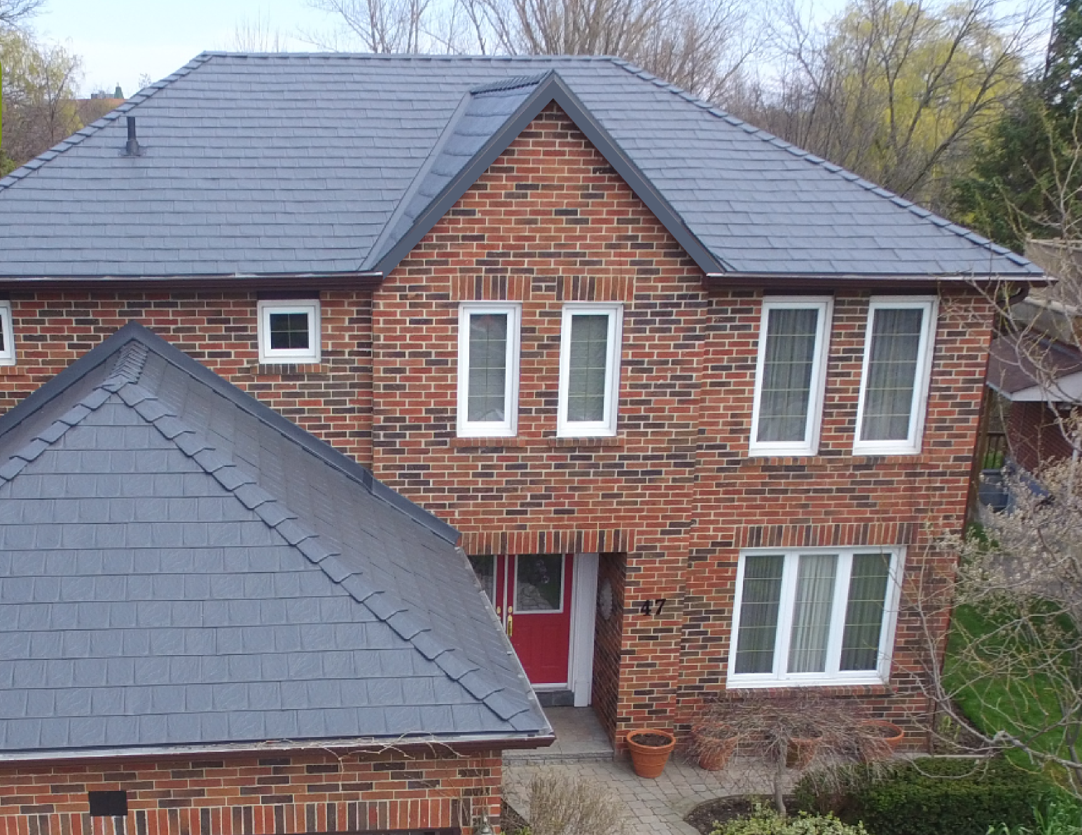 Maison à deux étages en briques rouges avec toiture en tuiles gris foncé, garage à deux portes, et porche d'entrée avec colonne blanche.