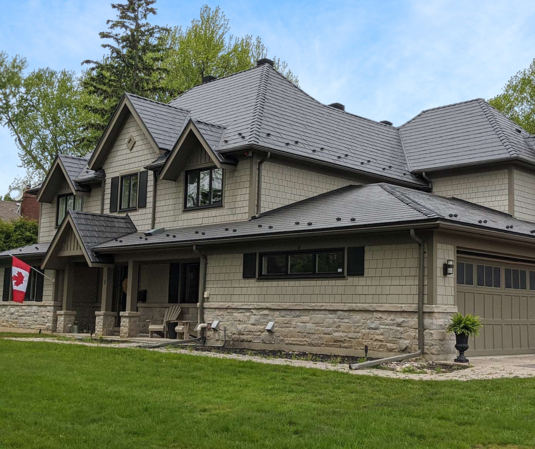 Une maison à deux étages avec une toiture en tuiles gris foncé, des fenêtres noires, un porche avec une banquette en bois, et un drapeau canadien dans le jardin. La maison possède un mélange de murs en pierre et en revêtement en bois, entourée d'un j