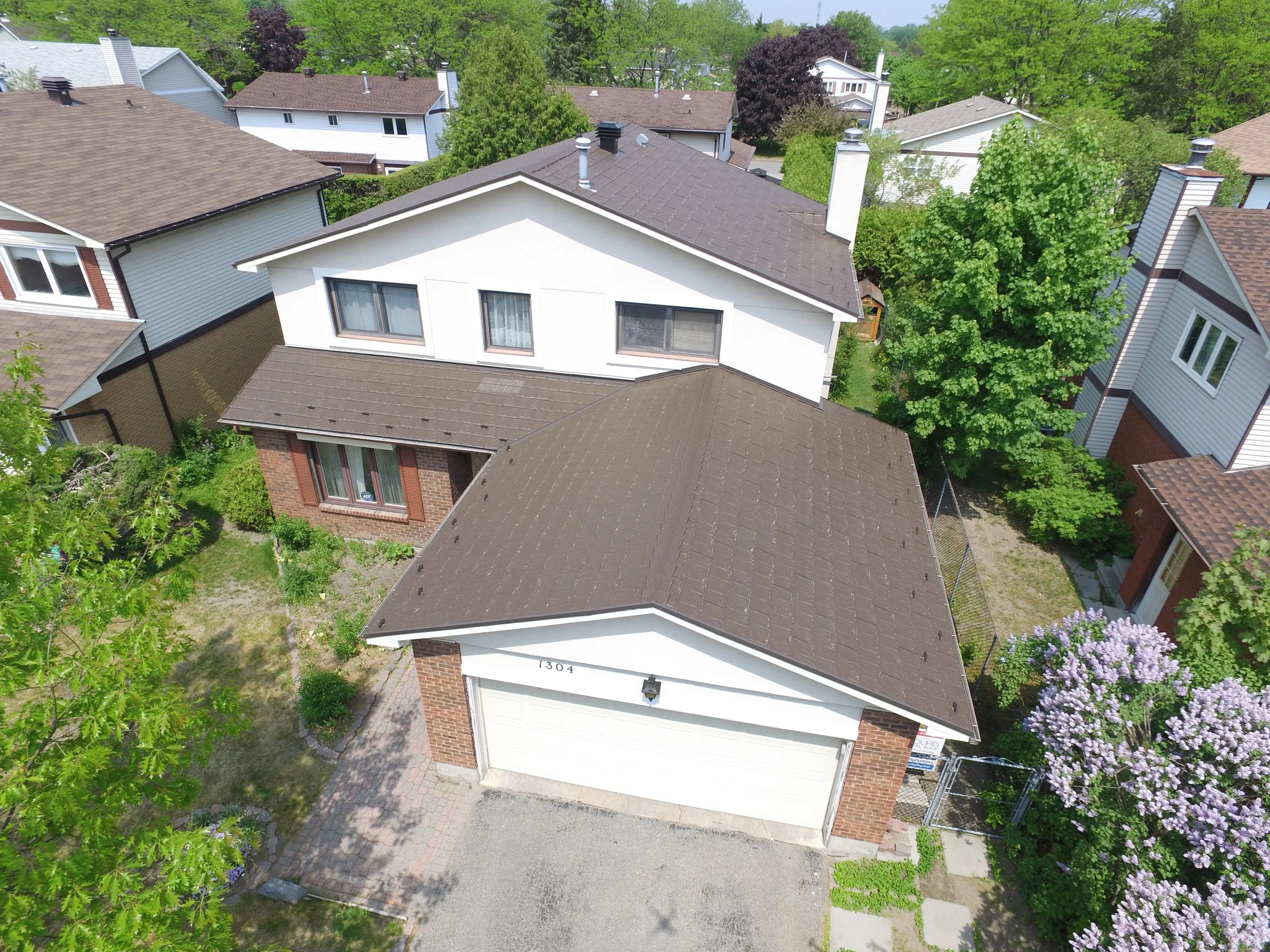 Vue aérienne d'une maison résidentielle avec un toit en bardeaux bruns, garage, entourée d'arbres et de maisons voisines dans un quartier résidentiel.
