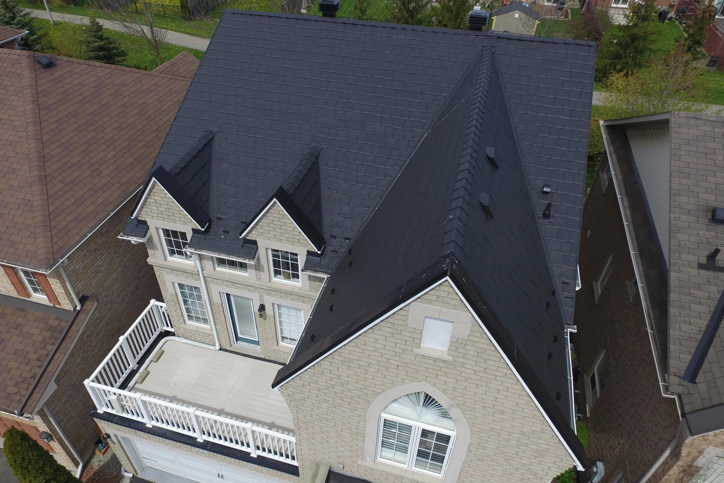 Vue aérienne d'une maison à deux étages avec un toit noir, un balcon blanc et des fenêtres à volets, entourée d'autres maisons avec différents styles de toiture.